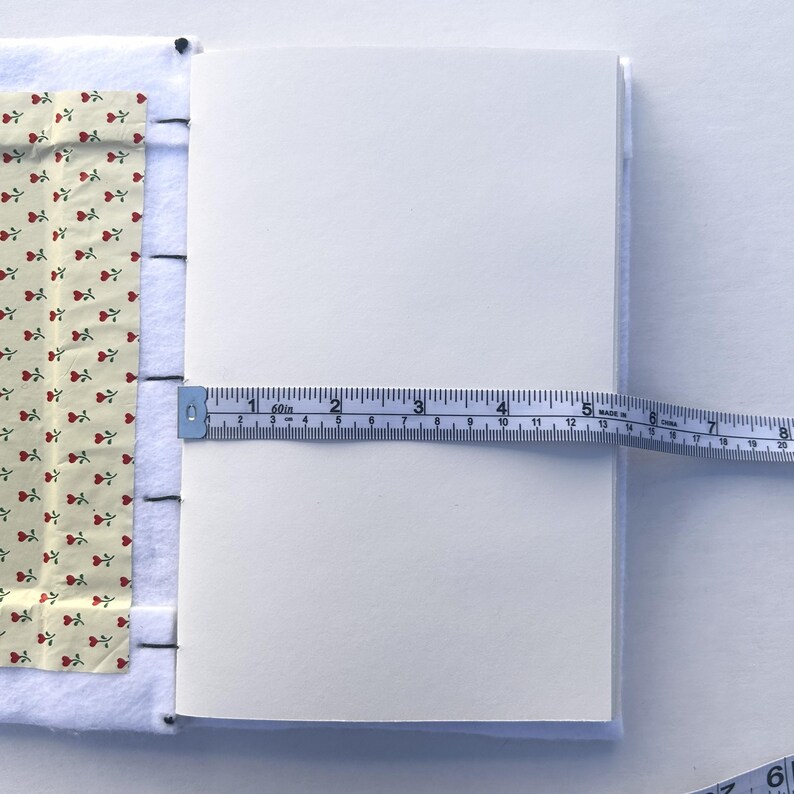 May include: A white notebook with a floral patterned fabric cover is measured with a blue measuring tape. The notebook is approximately 8 inches wide. The cover has a cream background with red rose patterns.
