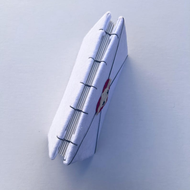 May include: A handmade white journal with a stitched binding. The cover features a red and white embroidered design. The pages are visible along the spine, and the journal is angled slightly, showcasing its handcrafted details.