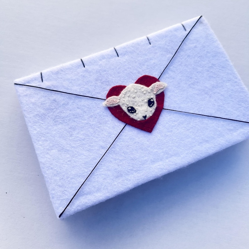 May include: A white felt envelope with black stitching and lines. A red heart with a white lamb applique is centered on the envelope. The lamb has small pink ears and black eyes.