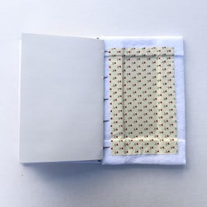May include: An open notebook with blank white pages. The right side features a cream-colored fabric with a repeating pattern of small red roses. The fabric is bordered by white felt and stitched to the notebook's spine.