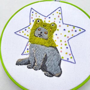 May include: Hand-stitched embroidery art featuring a gray cat wearing a lime green frog hat. The cat is centered on a white fabric, with a large star shape in the background, decorated with colorful dots. The embroidery is framed in a green hoop.