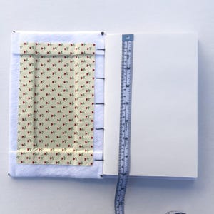 May include: Open notebook with white pages and a floral fabric cover featuring red roses on a cream background. A blue measuring tape shows the notebook's height. The notebook is approximately 6 inches tall.