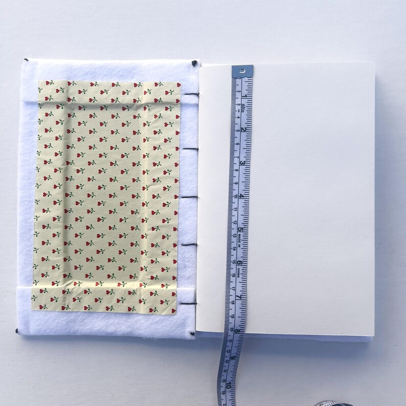 May include: Open notebook with white pages and a floral fabric cover featuring red roses on a cream background. A blue measuring tape shows the notebook's height. The notebook is approximately 6 inches tall.