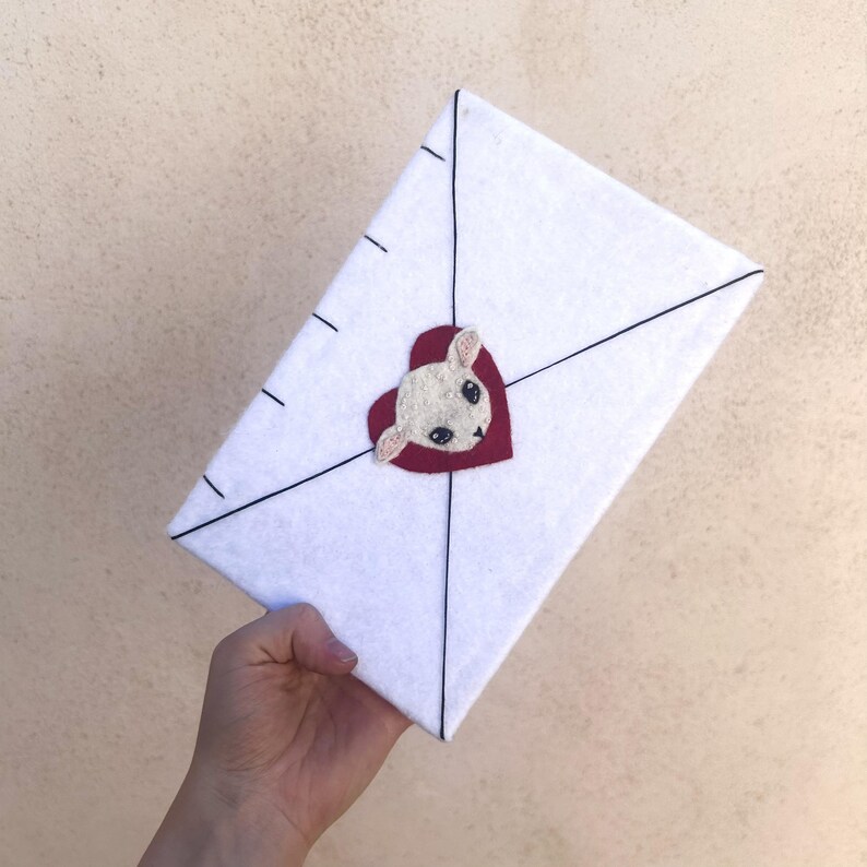 May include: A white felt envelope with black lines and dashes, featuring a red heart with a white bunny face. The envelope is held by a hand.