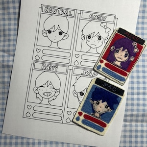 May include: Two felt patches with character portraits, one labeled "ANGRY" with a purple-haired character, and the other labeled "SAD" with a blue-haired character. A black and white drawing of four emotion faces is in the background.