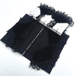 Pode incluir: Uma capa de livro de veludo preto com babados de renda preta nos bordos e um laço central preto. O interior apresenta páginas brancas encadernadas com costuras visíveis, criando um contraste entre o tecido escuro e o papel.