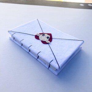 May include: A white felt journal shaped like an envelope, with black stitching and a red heart-shaped applique featuring a cream-colored sheep. The journal is closed with black thread.