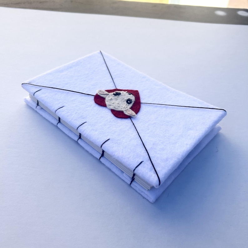 May include: A white felt journal shaped like an envelope, with black stitching and a red heart-shaped applique featuring a cream-colored sheep. The journal is closed with black thread.