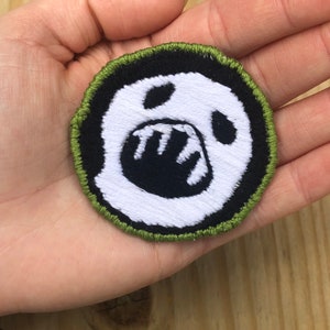 May include: A white embroidered patch with a black outline and a black and white image of a ghost with a wide open mouth and teeth. The patch has a green border.