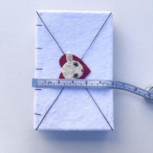 May include: A white felt envelope with a red heart and a bunny applique. The envelope is measured with a tape measure, showing a length of approximately 5 inches.