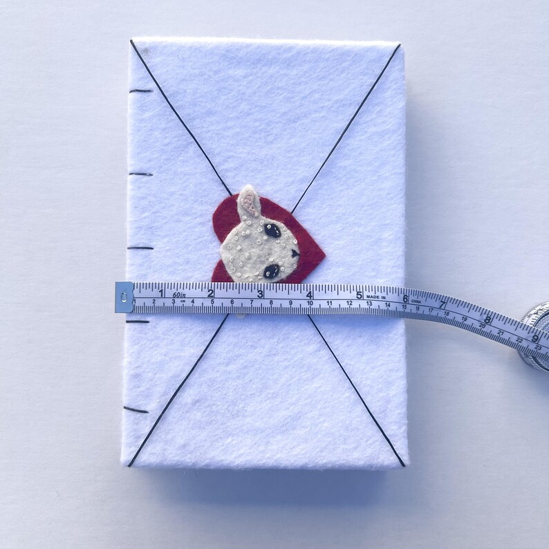 May include: A white felt envelope with a red heart and a bunny applique. The envelope is measured with a tape measure, showing a length of approximately 5 inches.