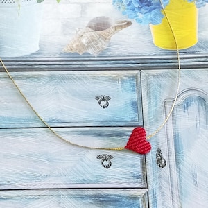 Puede incluir: Un collar con un colgante en forma de corazón rojo en una cadena dorada. El collar se muestra sobre una superficie de madera azul y blanca.