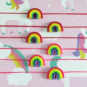 May include: Six rainbow-coloured bracelets with red string attached. The bracelets are made of colourful yarn and are shaped like rainbows. The bracelets are on a pink background with a unicorn pattern.