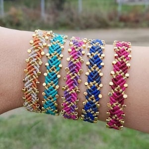 May include: Five colorful friendship bracelets with gold beads. The bracelets are woven with a chevron pattern and are stacked on a person's wrist.