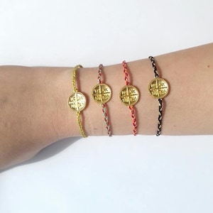 Op de afbeelding: Vier gevlochten armbandjes van touw met gouden bedels. Elke bedel heeft een kruisontwerp met opschrift. De armbandjes zijn in geel, blauw, rood en zwart.
