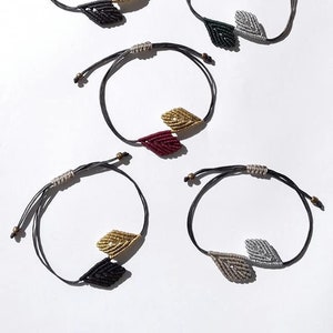 Könnte beinhalten: Fünf graue Armbänder aus Schnur mit verschiedenen farbigen Blatt-Anhängern. Die Anhänger sind aus geflochtener Schnur und in den Farben Gold, Grün, Silber und Rot.