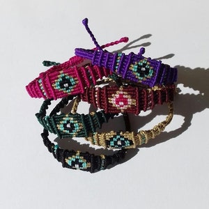 May include: Five colorful friendship bracelets with an eye design in the center. The bracelets are made with different colored threads, including pink, purple, green, gold, and black.