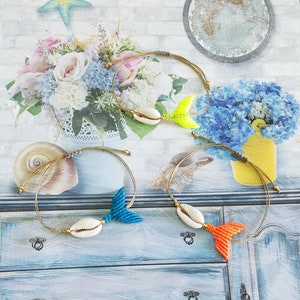 Può includere: Tre braccialetti dell'amicizia fatti a mano con un ciondolo a conchiglia e una coda di sirena in macramé colorata. I braccialetti sono realizzati con un cordoncino marrone e presentano una coda blu, gialla e arancione.