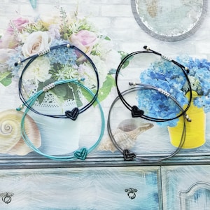 Könnte beinhalten: Drei Freundschaftsarmbänder mit schwarzen Herzanhängern. Die Armbänder sind aus blauem, türkisfarbenem und grauem Band gefertigt.