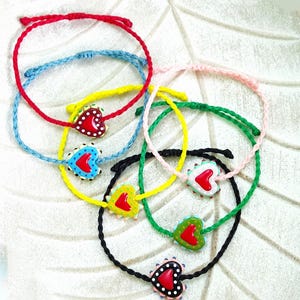 May include: A collection of colorful friendship bracelets. Each bracelet features a braided cord in red, blue, yellow, green, pink, and black. Each bracelet has a heart-shaped charm with a red heart and white polka dots.