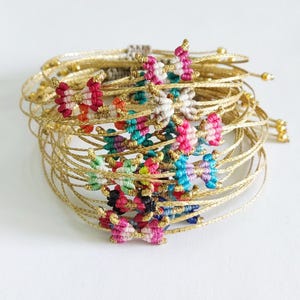 Puede incluir: Una pila de pulseras de alambre dorado con abalorios de mariposa anudados de colores. Las pulseras están apiladas una encima de la otra, con los abalorios de mariposa mirando hacia afuera.