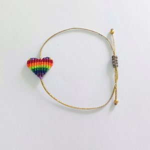 May include: A handmade bracelet featuring a rainbow-colored heart charm. The heart is made of woven threads in various colors. The bracelet has a gold-colored cord with adjustable knots and small gold beads. The background is plain white.