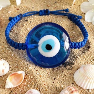 Könnte beinhalten: Ein blaues Nazar-Armband mit einem runden Glasanhänger, der konzentrische Ringe in Dunkelblau, Weiß und Hellblau sowie eine schwarze Pupille in der Mitte aufweist. Der Anhänger ist an einem geflochtenen blauen Band mit Schiebeknoten befestigt.