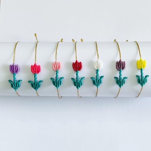 Op de afbeelding: Een verzameling handgemaakte macramé-armbanden, elk met een kleurrijk tulpontwerp. De tulpen zijn er in verschillende kleuren, waaronder paars, roze, rood, wit en geel, met groene stelen en bladeren. De armbanden worden op een witte ondergrond gepresenteerd.