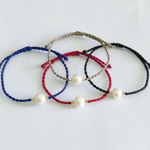 May include: Four braided string bracelets with a single pearl charm on each. The bracelets are in navy blue, red, black, and beige.