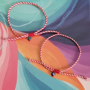 Può includere: Due braccialetti intrecciati rossi e bianchi con un piccolo ciondolo a forma di occhio blu e bianco. I braccialetti sono realizzati con un cordoncino ritorto e sono esposti su uno sfondo colorato con sfumature di blu, rosa e viola.