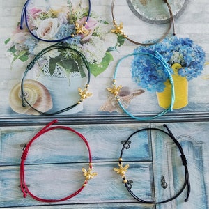 May include: Six colorful string bracelets with gold bee charms. The bracelets are displayed on a wooden surface with a floral background.