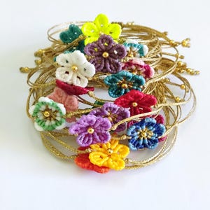 Puede incluir: Una colección de pulseras hechas a mano con diseños florales coloridos. Cada pulsera tiene un cordón dorado y una flor en tonos verdes, amarillos, morados, rojos, azules y blancos. Las flores tienen un centro de cuentas doradas.