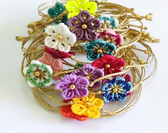 May include: A collection of handmade bracelets featuring colorful flower designs. Each bracelet has a gold-colored cord and a flower in shades of green, yellow, purple, red, blue, and white. The flowers have a gold bead center.