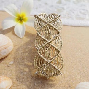 Peut inclure: Un bracelet en macramé doré au design complexe, posé sur une plage de sable. Une fleur blanche au centre jaune et des coquillages sont à proximité. L'océan est visible en arrière-plan.