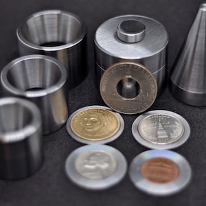 Puede incluir: Un conjunto de herramientas metálicas para hacer monedas, que incluye tres anillos de diferentes tamaños, un cilindro con un pequeño disco de metal en la parte superior y una herramienta en forma de cono. También hay cinco monedas de diferentes tamaños y diseños.