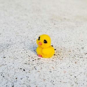 May include: A small, yellow chick figurine with black eyes and a pink beak and feet. The chick has a fluffy texture and is sitting on a speckled surface. The figurine is a cute, decorative item.