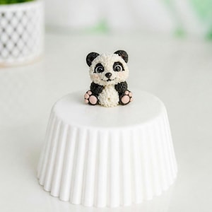 Puede incluir: Una pequeña figura de panda, en blanco y negro, con patas rosas, posada sobre un pedestal blanco acanalado. El panda tiene una cara sonriente. La figura es un artículo decorativo.