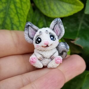 Puede incluir: Una pequeña figurita de chinchilla pintada a mano, de color blanco con orejas grises y una nariz rosa. La figurita está sentada en una mano con follaje verde en el fondo.