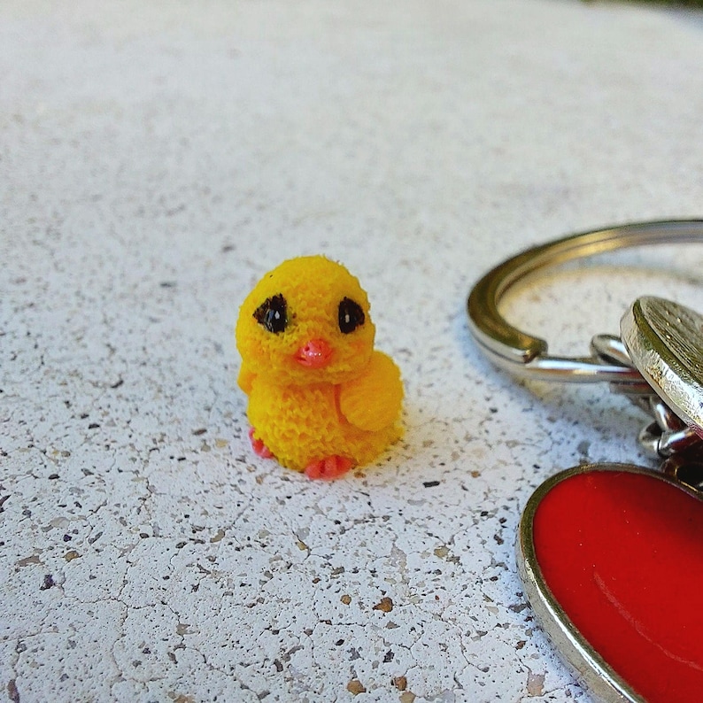 May include: A small, yellow chick figurine with black eyes and an orange beak. The chick has a textured surface and is sitting on a white surface. A silver keychain with a red heart charm is in the background.