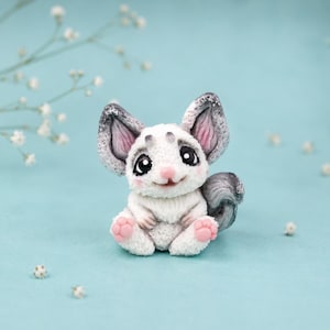 Può includere: Una piccola statuetta scolpita di un cincillà con pelliccia bianca, grandi occhi scuri e accenti rosa su naso, orecchie e cuscinetti delle zampe. La creatura ha orecchie con punte grigie e una coda grigia, seduta in posizione eretta e sorridente.