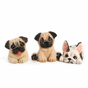 May include: Three small dog figurines made of resin. On the left, a tan pug with a black face and a red tongue sticking out. In the center, a tan pug sitting upright with a black face. On the right, a white and black French bulldog lying down.