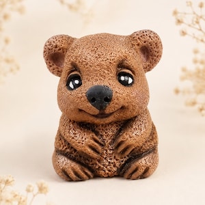 May include: A small, brown, textured figurine of a quokka with rounded ears, large dark eyes, and a black nose. The creature is depicted in a seated position with its front paws resting together near its chest, featuring a subtle, smiling expression.
