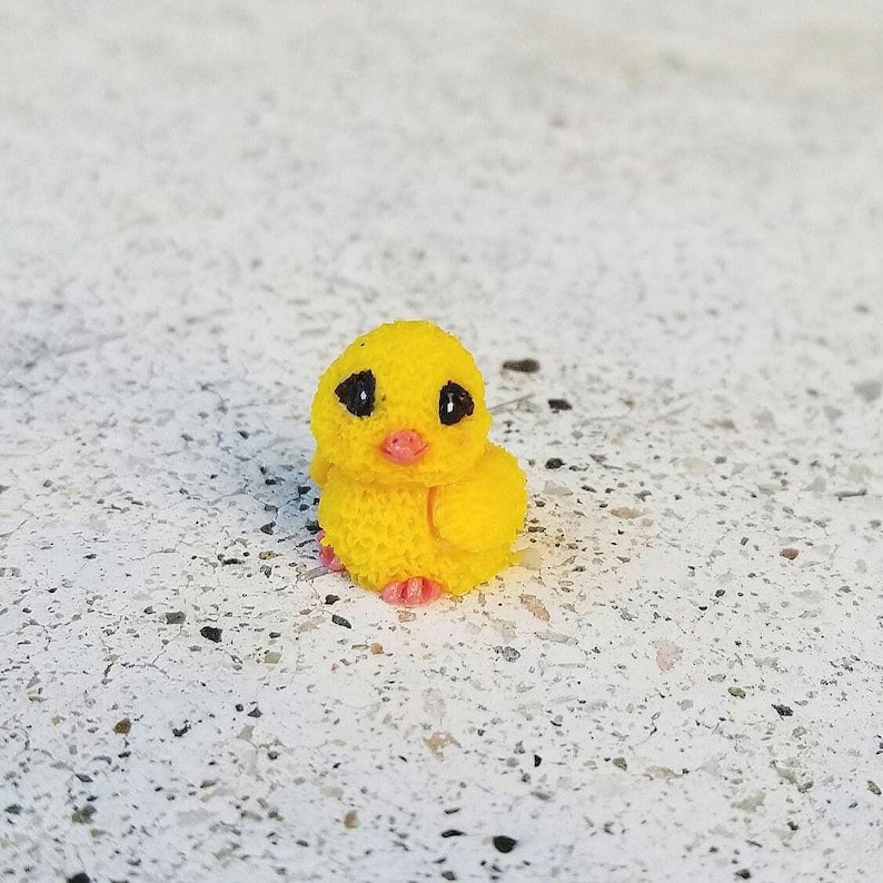 May include: A small, yellow chick figurine with black eyes and a pink beak and feet. The chick has a textured, fluffy appearance and is sitting upright. The background is a speckled, light-colored surface.