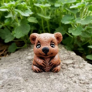 May include: A small, brown, textured figurine of a quokka with large, dark, glossy eyes and a black nose. The creature is depicted in a seated position with its front paws resting against its chest, featuring a smooth, rounded head and ears.