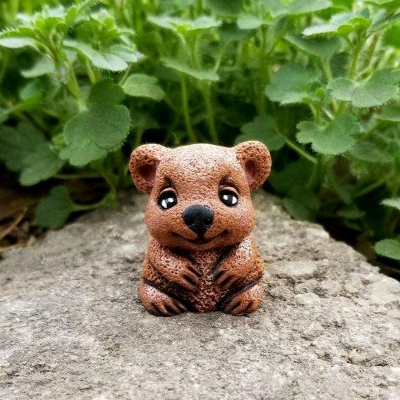 May include: A small, brown, textured figurine of a quokka with large, dark, glossy eyes and a black nose. The creature is depicted in a seated position with its front paws resting against its chest, featuring a smooth, rounded head and ears.