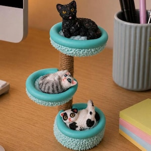 Könnte beinhalten: Eine dreistufige Katzenbaum-Figur mit drei Miniatur-Katzenfiguren. Der Katzenbaum hat einen mit Seil umwickelten Pfosten und türkisfarbene Betten mit flauschigem Rand. Die Katzen sind schwarz, grau und weiß sowie schwarz und weiß.
