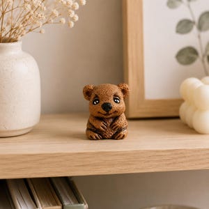May include: A small, brown, textured figurine of a quokka with large, dark, expressive eyes, a black nose, and a subtle smile. The creature is depicted in a seated position with its front paws resting against its chest, showcasing detailed fur texture.