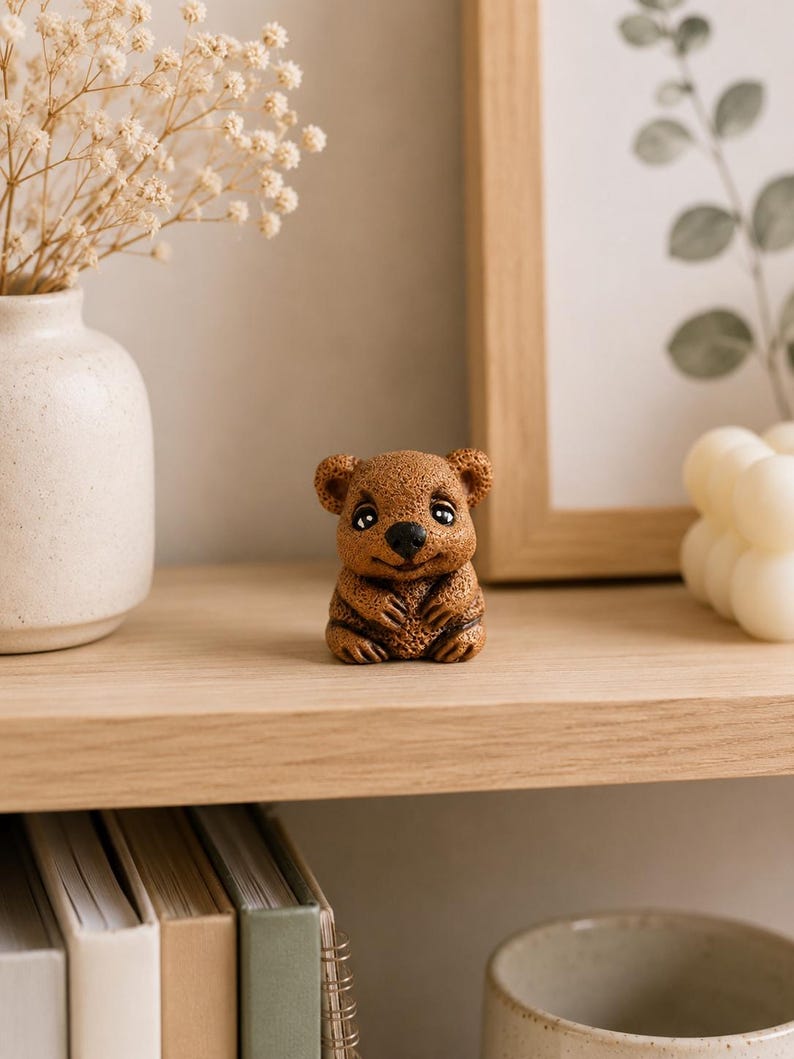 May include: A small, brown, textured figurine of a quokka with large, dark, expressive eyes, a black nose, and a subtle smile. The creature is depicted in a seated position with its front paws resting against its chest, showcasing detailed fur texture.