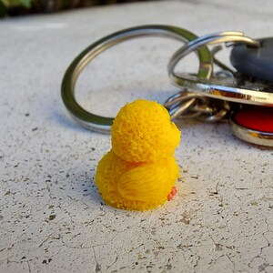 May include: A bright yellow rubber ducky keychain. The duck has a textured body and a small, pink foot. A silver key ring is attached to the duck, with keys visible in the background.