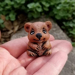 May include: A small, textured brown figurine of a quokka with rounded ears, large dark eyes, and a black nose. The creature is depicted in a seated position with its front paws resting together near its chest, featuring a smooth, matte finish.